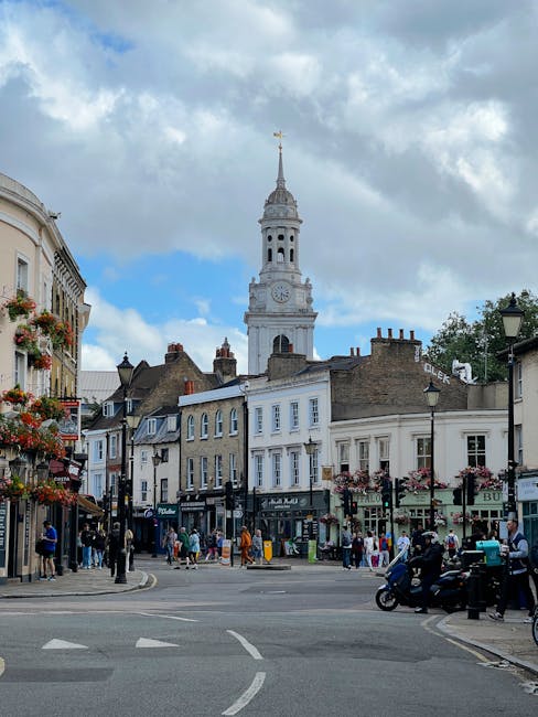 A busy street scene in Enfield town centre during daytime, featuring a variety of residential and commercial buildings, including shops and cafes, with pedestrians walking on the pavement and crossing the road. In the background, a tall, historic clock tower with a spire and weather vane stands prominently above the surrounding structures, partly obscured by a cloudy sky with patches of blue. On the street, there are a few vehicles, including motorcycles and cars, some waiting at the junction, while others are parked along the roadside. Street lamps and hanging flower baskets adorn the area, contributing to a vibrant and lively atmosphere typical of a popular town centre in London. This scene reflects an environment where professional removals by Movers Enfield might take place during a home relocation or furniture transport process, with visible surroundings conducive to moving logistics and packing activities.