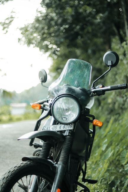 A close-up view of a black motorcycle parked on the side of a road during daytime. The motorcycle features a clear windscreen, round headlight, orange turn signals, and rearview mirrors connected to the handlebars. The front wheel and tire are visible, along with the front suspension system. The license plate is readable, displaying the registration number