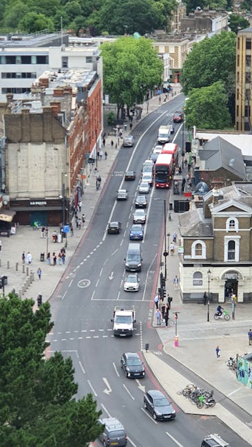 A busy street scene in Enfield town centre during daytime, featuring a variety of residential and commercial buildings, including shops and cafes, with pedestrians walking on the pavement and crossing the road. In the background, a tall, historic clock tower with a spire and weather vane stands prominently above the surrounding structures, partly obscured by a cloudy sky with patches of blue. On the street, there are a few vehicles, including motorcycles and cars, some waiting at the junction, while others are parked along the roadside. Street lamps and hanging flower baskets adorn the area, contributing to a vibrant and lively atmosphere typical of a popular town centre in London. This scene reflects an environment where professional removals by Movers Enfield might take place during a home relocation or furniture transport process, with visible surroundings conducive to moving logistics and packing activities.