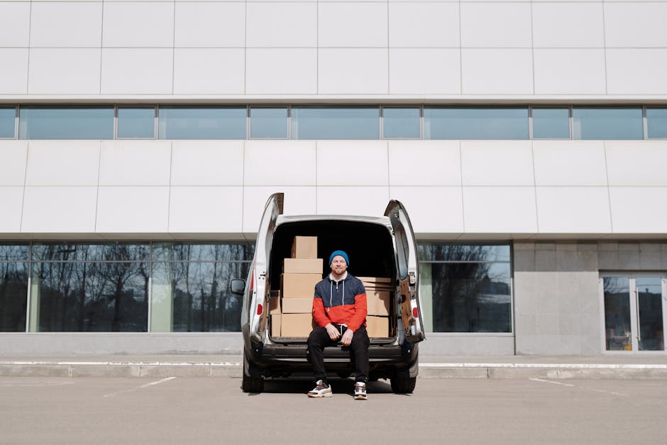 A man with a beard, wearing a red baseball cap, a checked shirt, and a two-tone brown and beige jacket, stands beside the open rear of a van during a house removal. Inside the van, there are several c