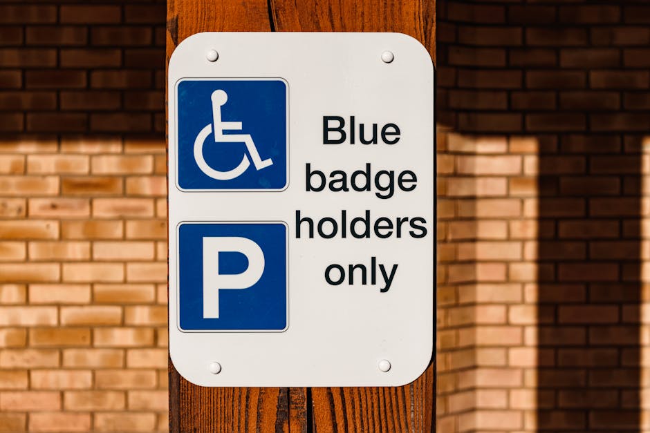 A white rectangular parking sign mounted on a wooden post beside a brick wall, featuring a blue wheelchair symbol indicating accessible parking and a blue 'P' symbol below, accompanied by the text 'Blue badge holders only'. The sign is photographed in natural daylight, with the shadow of the post visible on the brick wall. The image is relevant for house removals and moving services, such as those offered by Movers Enfield, especially when discussing parking permits or access regulations during home relocation and furniture transport processes.