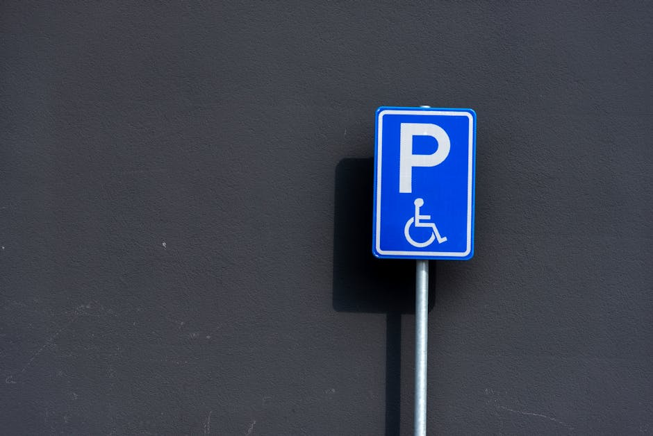 A blue and white parking sign indicating a designated space for wheelchair users, mounted on a metal post attached to a dark grey wall. The sign includes a large letter 'P' and a wheelchair accessibility symbol below it. The sign casts a shadow to the left due to lighting. In the background, part of the street or pavement is not visible, but the focus remains on the sign's clear visibility and position on the exterior property. The image supports content about house removals and property logistics, with Movers Enfield possibly referencing parking arrangements for home relocation and furniture transport services in Enfield.