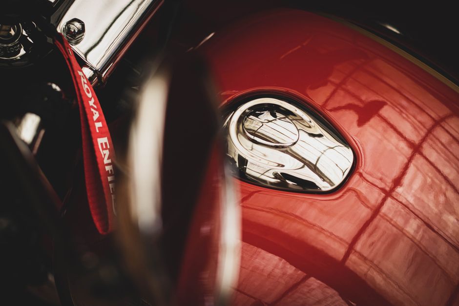 Close-up view of a red classic car's front hood, focusing on the shiny, rounded headlight reflecting the surrounding environment, including a silhouette of a person and structural elements like windows and beams. Part of the car's chrome bumper and hood ornament are visible, with the glossy surface showing subtle reflections of the interior space where the vehicle is parked. The scene is well-lit, highlighting the vehicle's smooth, polished paint and chrome detailing. The background hints at an indoor setting, possibly a garage or warehouse, with objects such as boxes, packing materials, and tools nearby, indicating a home relocation or vehicle transportation context. This detailed image captures the careful handling of vehicle transport during moving or packing and aligns with professional removals services offered by Movers Enfield, especially those involving specialist transport or packing of valuable or delicate items.