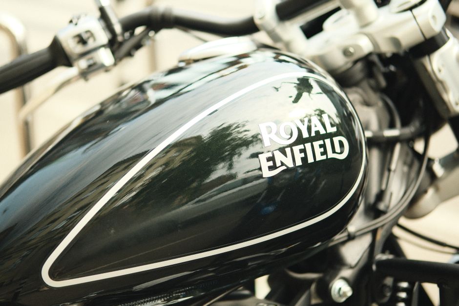 Close-up view of a black motorcycle fuel tank with a glossy finish, featuring the white logo 'Royal Enfield' prominently displayed on the side. The tank's surface reflects the surrounding environment, including trees and sky, indicating outdoor lighting. Visible parts include the fuel cap, surrounding metal frame, and some mechanical components beneath the tank. The motorcycle is situated in a setting that suggests either being prepared for transportation or part of a home relocation process, with other moving equipment or vehicles possibly nearby. The image emphasizes the details of the motorcycle's design, materials, and positioning within a moving or packing context relevant to house removals and furniture transport, as handled by Movers Enfield.