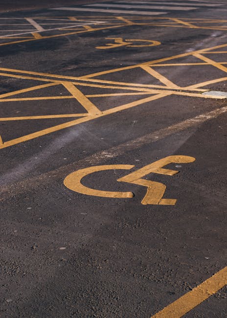 Close-up view of an outdoor parking area showing two designated accessible parking spaces marked with yellow wheelchair symbols painted on dark asphalt. The second space is partially visible behind th