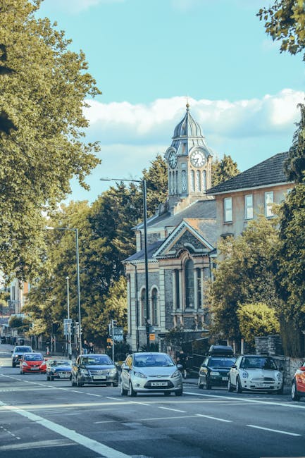 A daytime view of a busy street in Palmers Green with multiple parked and moving cars along the roadside. On the left side, there are a row of mature trees with green foliage partially shading the street and sidewalk. In the background, a historic church with a tall clock tower featuring a domed spire and a clock face is visible, surrounded by additional buildings and greenery. The scene captures the typical urban environment suitable for house removals or home relocation services. The image highlights the infrastructure and environment where Movers Enfield offer same day removal and furniture transport, emphasizing the importance of efficient loading and moving logistics in this residential area.